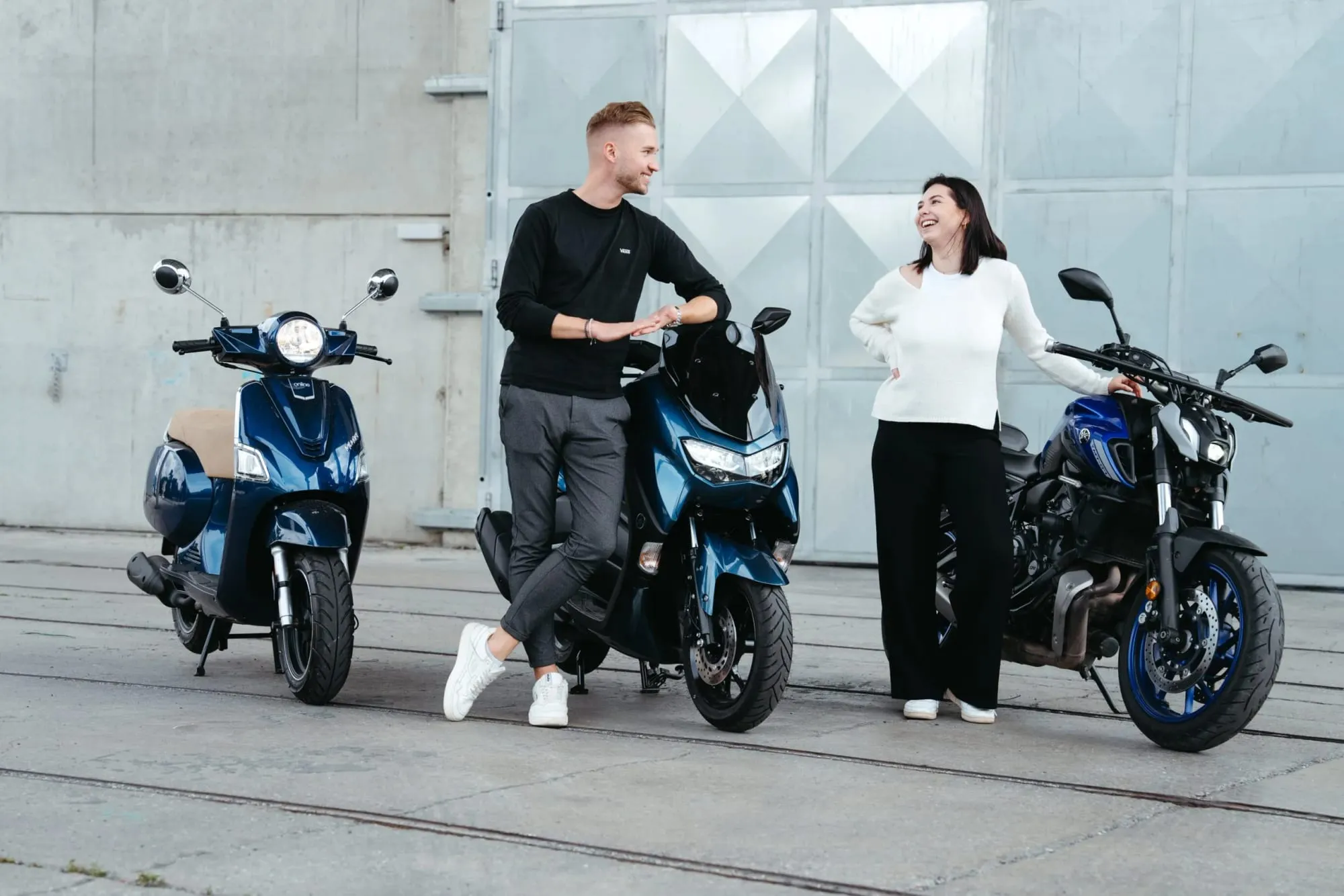 Zwei junge Leute lehnen lachend an einem Motorroller und einem Motorrad
